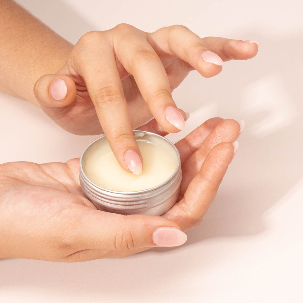 Doigts appliquant une crème réparatrice contenue dans un pot métallique, texture riche et onctueuse, illustrant un soin concentré en actifs végétaux pour la peau.